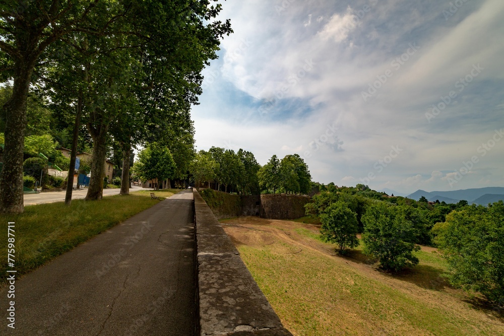 Concrete sidewalk surrounded by lush green trees under blue cloudy sky in Bergamo, Italy