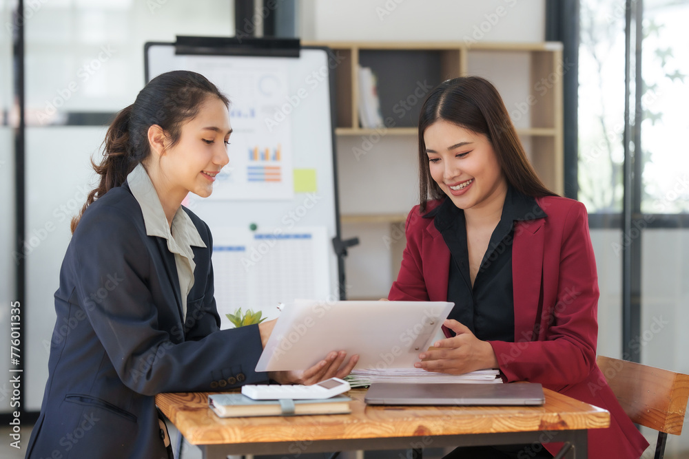 Two asian businesswoman in suits are sitting at a table with a white board behind them Two asian businesswoman in suits are sitting at a table with a white board behind them