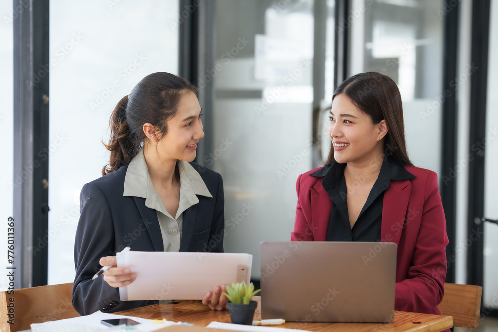 Two asian businesswomen in suits are sitting at a table with a laptop and a piece of paper Two asian businesswomen in suits are sitting at a table with a laptop and a piece of paper