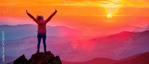 A woman, in celebration of reaching the summit of a mountain peak at sunset or sunrise, holds her arms high above her head in elation.