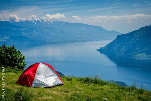 A red and white tent on the grassy hill overlooking the lake in Switzerland with mountains in the background. A view from the top of a mountain at summer time