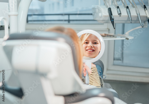 Little girl sitting in stomatology clinic chair and smiling at mirror showing her teeth after teeth dental procedures. Healthcare, kid's health and medicare industry concept image