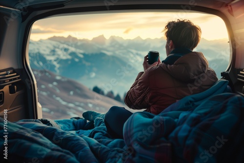 A man sitting in the trunk of his car, holding coffee and looking at mountains through an open window with blue blankets on him, camping gear inside, sunset light photo