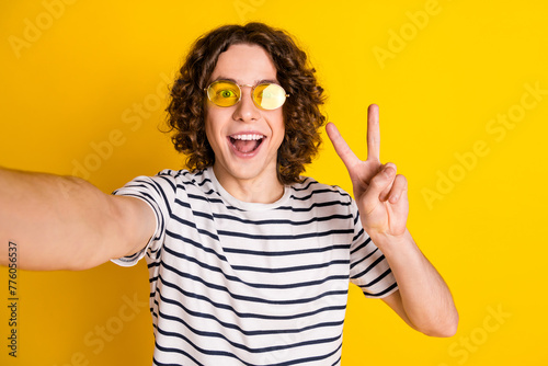 Photo portrait of handsome teenager guy take selfie show v-sign dressed stylish striped garment isolated on yellow color background