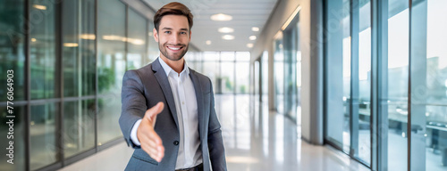 Confident businessman extending a handshake in a modern office corridor. Welcoming gesture from a smiling professional in a well-lit, contemporary workspace.