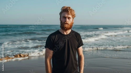 Light-skinned male model with a beard standing by the shore, wearing a black T-shirt mockup photo