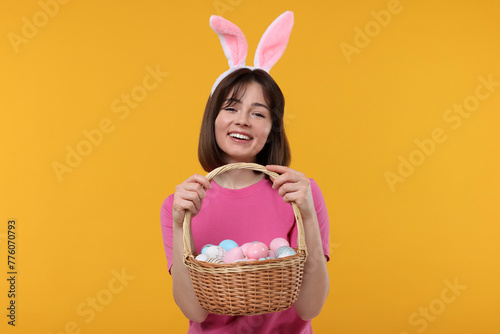 Easter celebration. Happy woman with bunny ears and wicker basket full of painted eggs on orange background