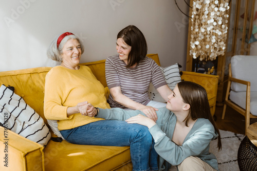 Happy senior woman spending leisure time with granddaughter and daughter-in-law while sitting on sofa at home