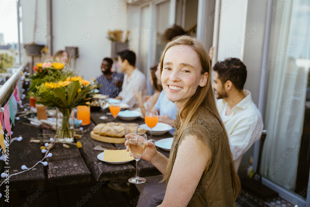 Side view of smiling woman holding wineglass while sitting with friends at dining table during dinner party in balcony Side view of smiling woman holding wineglass while sitting with friends at dining table during dinner party in balcony