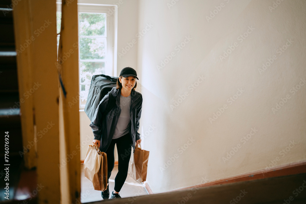 Smiling female delivery person holding bags and moving up on building staircase Smiling female delivery person holding bags and moving up on building staircase