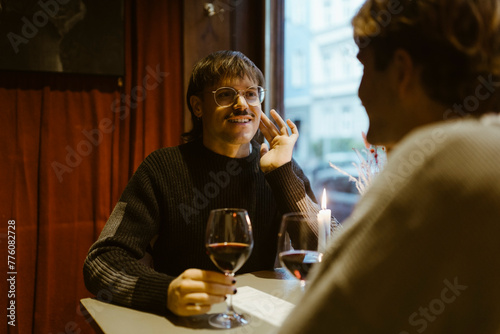Smiling gay man holding wineglass and talking with boyfriend while sitting at restaurant photo