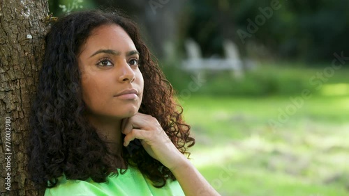 Beautiful thoughtful mixed race African American girl biracial teenager young woman outside thinking, sad or depressed sitting leaning against a tree, slow motion 4K clip
