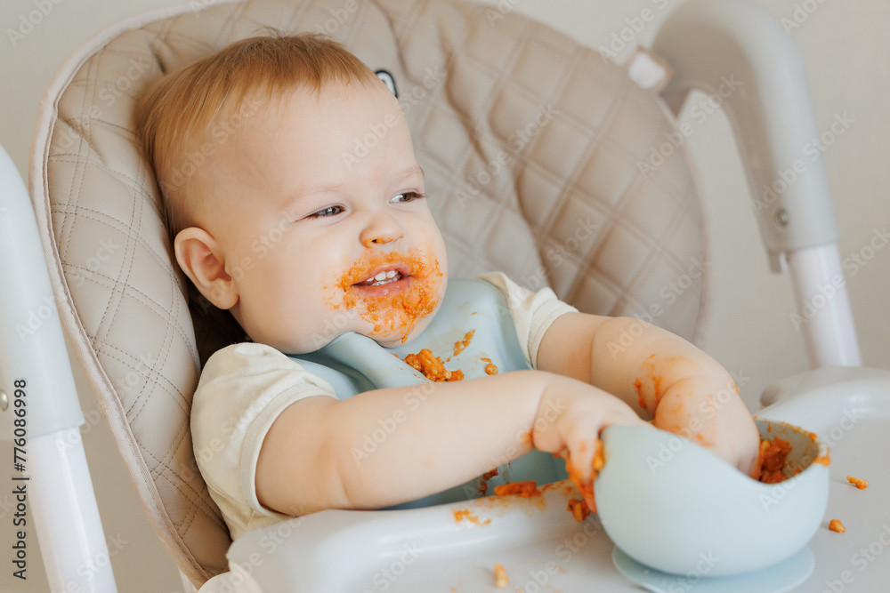 Happy messy baby boy eating pasta with tomato enjoys food sits In high chair, light background Happy messy baby boy eating pasta with tomato enjoys food sits In high chair, light background