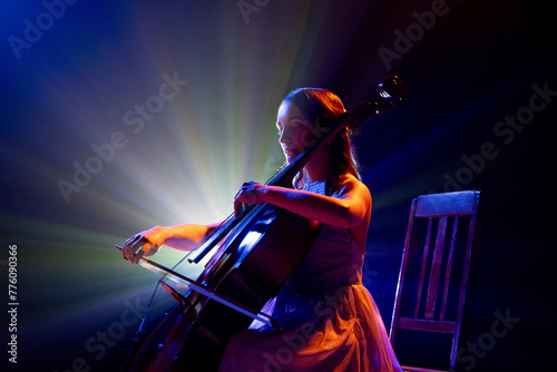 Talented young woman, cellist immersed in music, with striking halo of stage lights accentuating her silhouette on stage. Concept of hobby and work, music festivals, concerts, symphony show, culture.