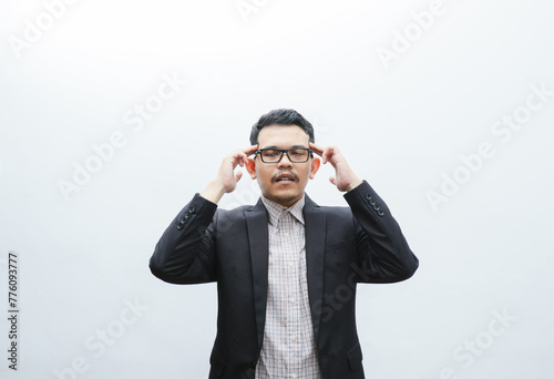 Tensed and distressed asian young man in black suit suffering big headache, close eyes, frowning and grimacing from pain, rubbing temples, have headache, need painkillers, white background © IgnatiusHarly