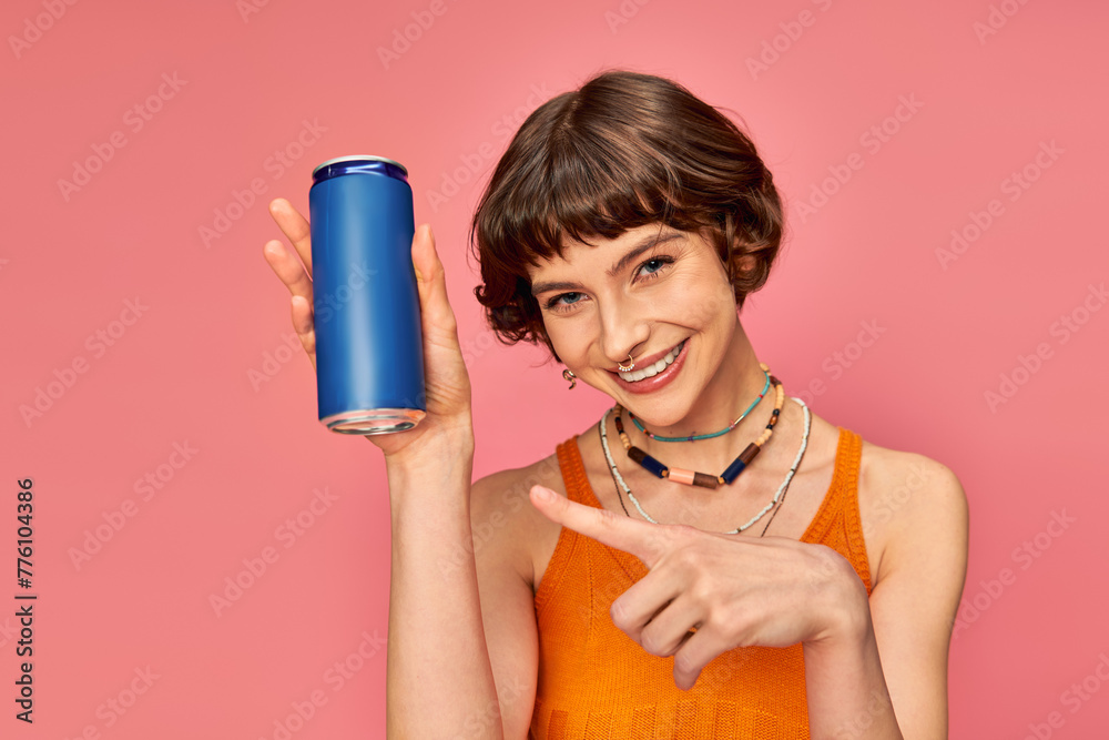 joyful young woman with short brunette hair pointing at soda can on pink, summer beverage