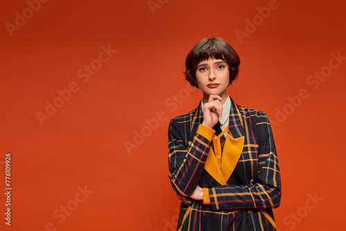thoughtful college girl with short hair posing in checkered uniform on orange background, decision