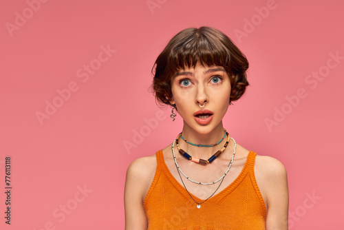 astonished young girl in her 20s standing in orange knitted tank top on pink background, disbelief