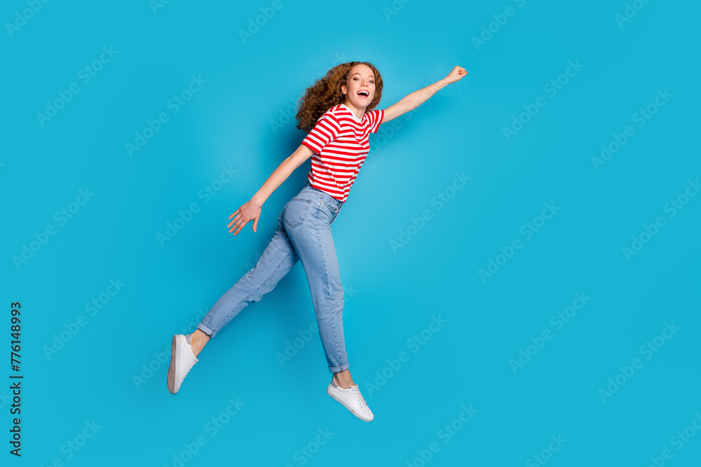 Full size photo of overjoyed girl wear stylish t-shirt pants jump stretching fist to empty space isolated on blue color background