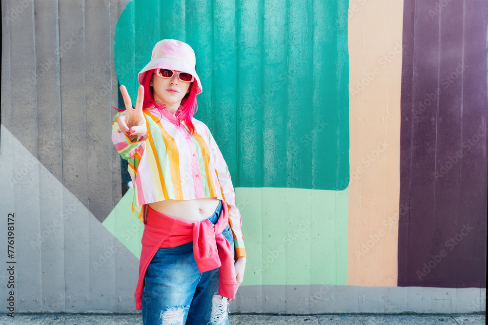 Young woman with pink hair and sunglasses in Bucket hat and multicolor strippled shirt making V sign by fingers on graffiti wall background. Street artist, hipster, blogger. Urban street fashion - obrazy, fototapety, plakaty 