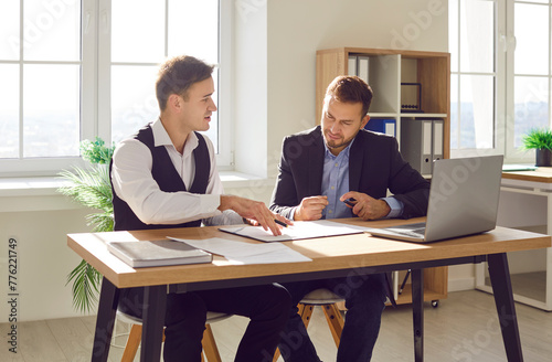 Two young confident company employees wearing suits sitting at the desk on workplace working in office having discussion, making a deal or reaching agreement going to sign a contract.