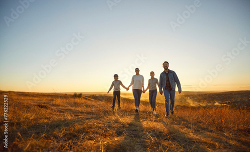 Happy smiling family of four with two kids boy in pilot's glasses and girl walking in the field together holding hands enjoying time together in nature at sunset time. Family leisure outdoors concept