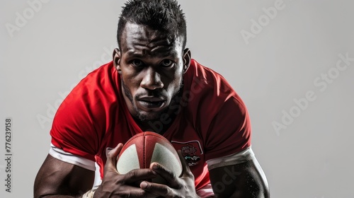 A male rugby player in a red jersey is holding a rugby ball in his hands, looking directly into the camera.