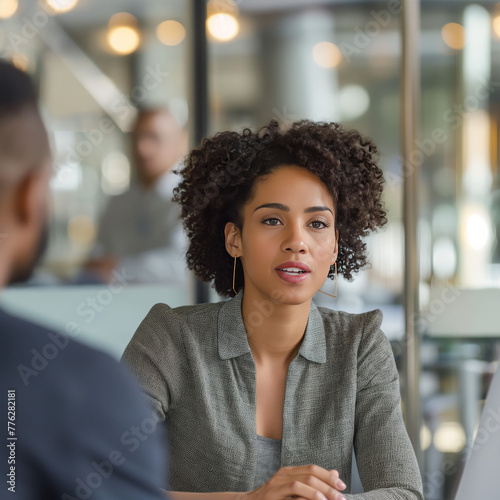 an african american woman interviews a hispanic male for a job. They are both dressed in modern business casual. They are in a conference room with glass walls that show a busy open office environment