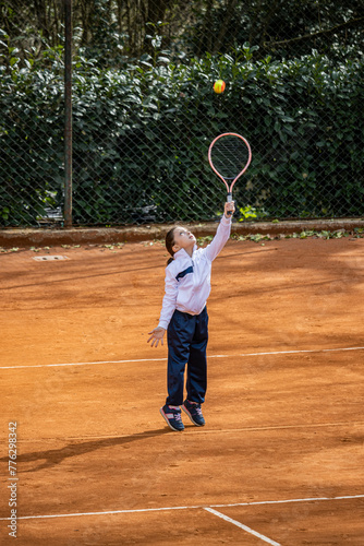 A little girl plays tennis on a red clay court. She runs and trains to learn the beautiful sport of tennis. Competitiveness, athlete, sporty child. © Ragemax