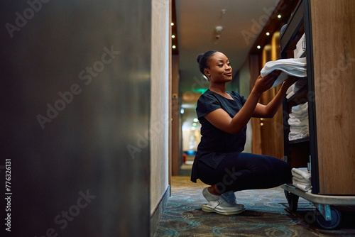 Young black maid taking clean towels from  cart while working in  hotel. photo
