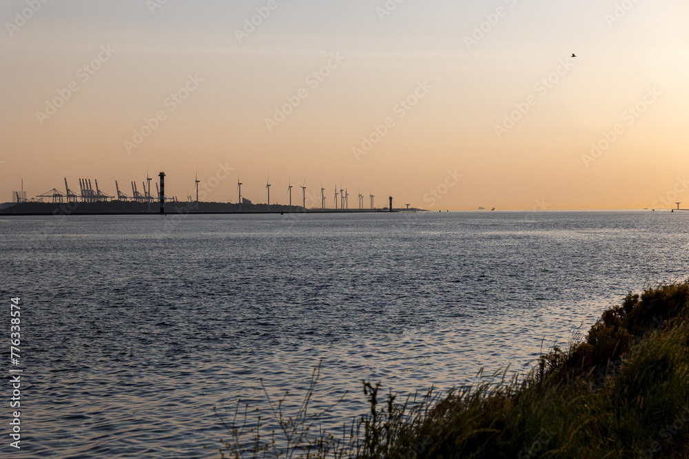 Beautiful windmills in the port of Rotterdam. Beautiful sunset on the sea coast. The Blue North Sea and Water surface. The lighthouse and shore are lit by the sunset sun.
