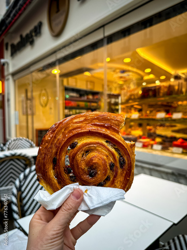 Photography Hand holding freshly baked pain aux raisin in front of  artisan shop “Maison Thevenin” at Rue de Buci in Paris