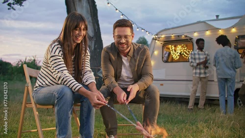 Positive woman telling funny history to her loving man during frying sausages on bonfire. Group of multi-ethnic friends communicating next to trailer with glowing garland. Campsite outdoors. Weekend.