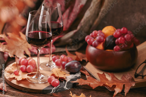 Autumn still life with two glasses of red wine, grapes, book and dry leaves in rustic style on dark wooden background. Romantic sweater weather concept with copy space