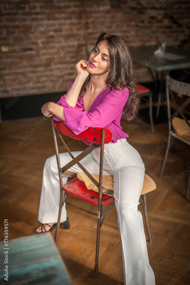 Beautiful young brunette woman in a purple blouse and white pants posing indoor against a brick wall. Portrait of a beautiful young brunette woman