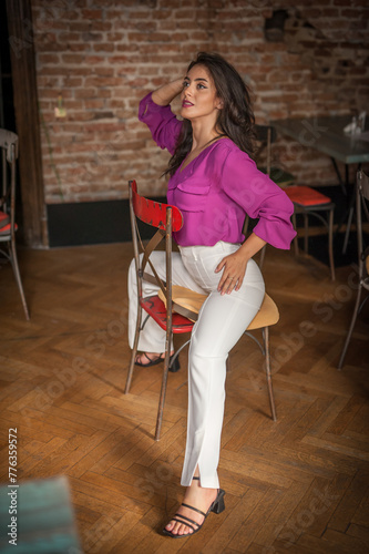 Obraz na płótnie Beautiful young brunette woman in a purple blouse and white pants posing indoor against a brick wall. Portrait of a beautiful young brunette woman w salonie