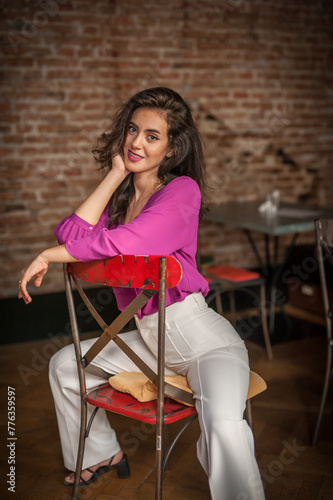 Obraz na płótnie Beautiful young brunette woman in a purple blouse and white pants posing indoor against a brick wall. Portrait of a beautiful young brunette woman w salonie