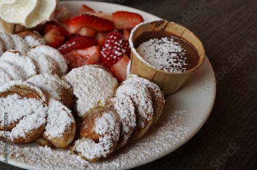 a plate of poffertjes, a traditional Dutch pancake, covered in sugar powder with a side of strawberry fruit and chocolate sauce 