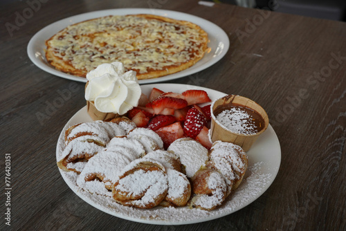 a plate of poffertjes, a traditional Dutch pancake, covered in sugar powder with a side of strawberry fruit and chocolate sauce. A savory pancake with a cheese, bacon, and egg in the background
