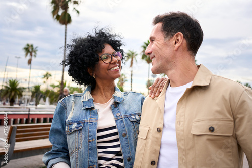 Smiling mature multiracial tourist couple embraced look at each other in love enjoying together free time on weekend on street in tourism city. Middle-aged husband and wife in vacation spring day #776458144