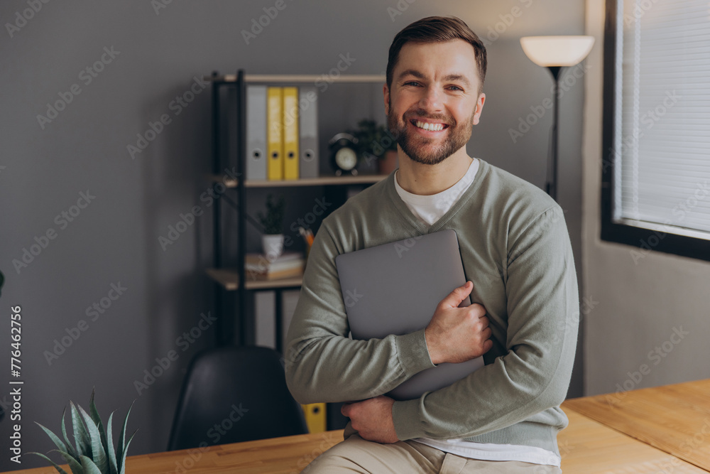 Modern happy bearded office male worker of IT company smiling and holding laptop in office