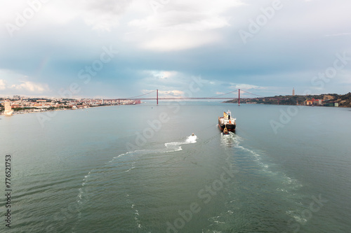 Aerial drone shot of cargo ship moving in Tagus river with Ponte 25 de Abril in background. The iconic Ponte 25 de Abril spans the background, set against the backdrop of a cloudy sky.