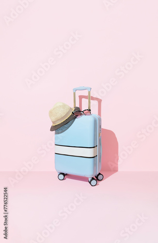 Blue suitcase with sunglasses and hat on a pastel pink background with long deep shadows. Travel concept