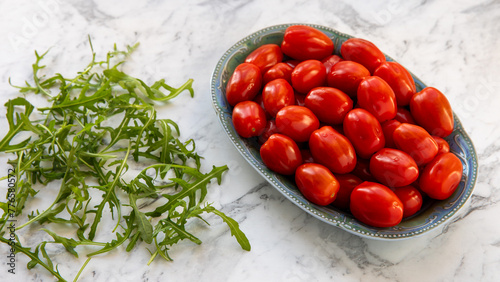 Whole, ripe date tomatoes on a plate and some rocket leaves on the side. Gray marble background.