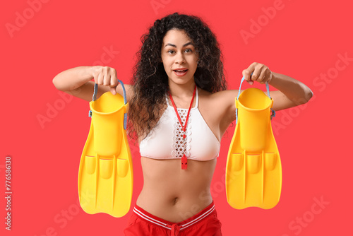 Beautiful young shocked African-American female lifeguard with flippers on red background
