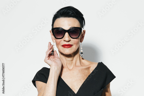 Elegant older woman in black dress and sunglasses posing in front of white wall portrait © SHOTPRIME STUDIO