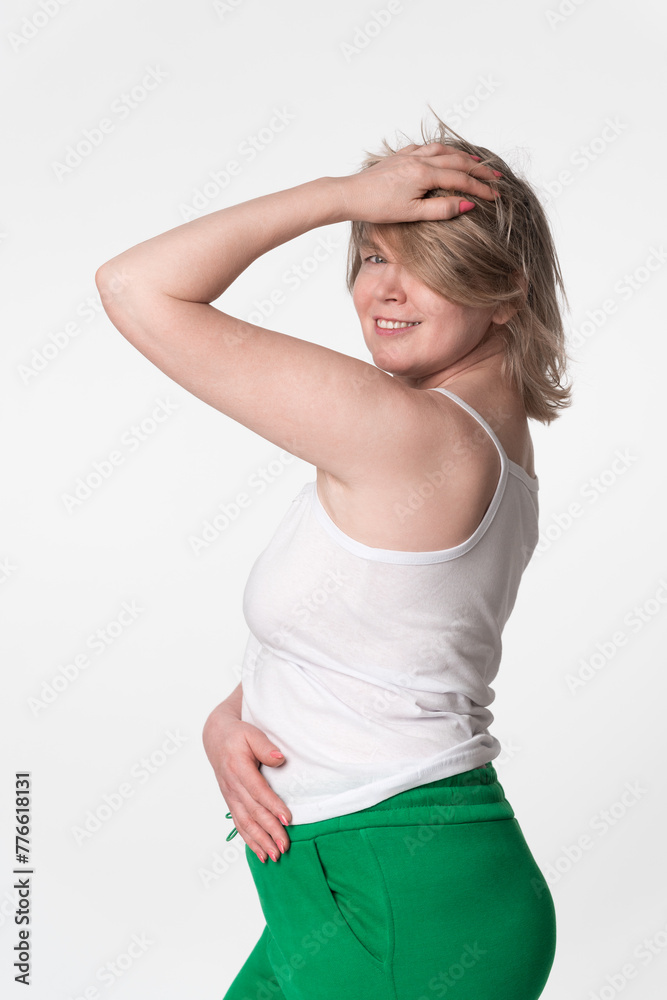 Portrait of happy plump woman is smiling and looking over her shoulder at camera, posing with one hand raised to head and hand on stomach. She wearing camisole and green sweatpants on white background Portrait of happy plump woman is smiling and looking over her shoulder at camera, posing with one hand raised to head and hand on stomach. She wearing camisole and green sweatpants on white background