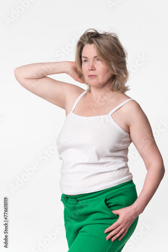 Portrait of chubby mature adult blonde woman Generation X model wearing cotton camisole and green jogging pants. Comely woman with one hand raised behind head looking away, posing on white background © Alexander Piragis