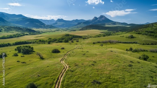 Aerial perspective of a hiking trail winding through a lush green meadow, leading towards a breathtaking mountain range