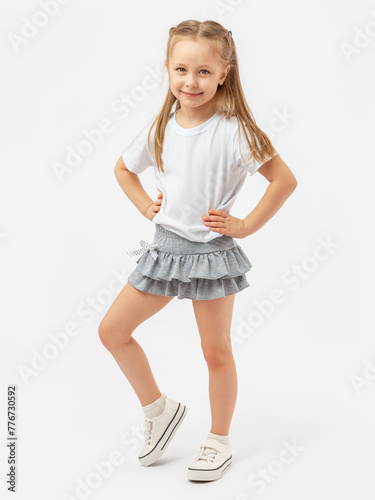 Sports dancing, children's aerobics. A 5-year-old girl stands smiling in a white T-shirt , sneakers and a gray skirt on a white background. Hands on the waist. Photo. Copy space.